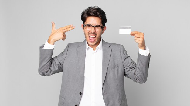 Young hispanic man looking unhappy and stressed, suicide gesture making gun sign and holding a credit card