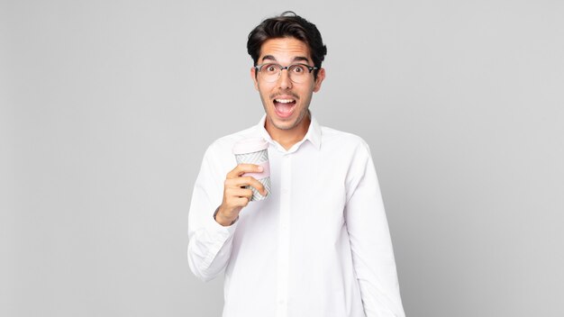 Young hispanic man looking very shocked or surprised and holding a take away coffee