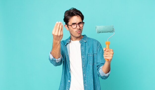 Young hispanic man making capice or money gesture, telling you to pay and holding a paint roller