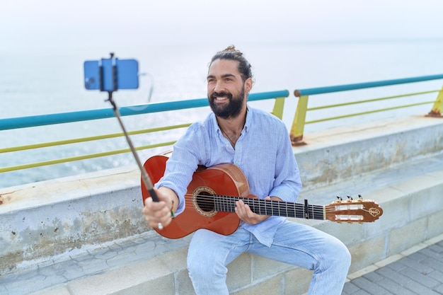 Young hispanic man musician holding classical guitar having video call at seaside