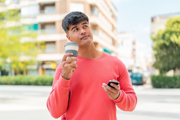 Молодой латиноамериканец на улице пользуется мобильным телефоном и держит кофе