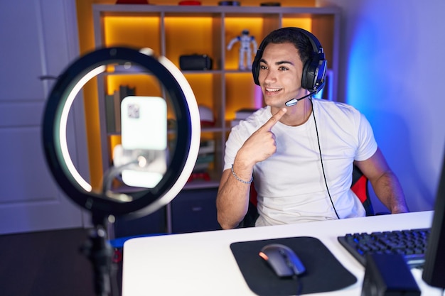 young hispanic man playing video games recording with smartphone smiling happy pointing with hand and finger
