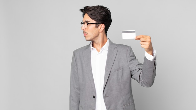 Young hispanic man on profile view thinking, imagining or daydreaming and holding a credit card