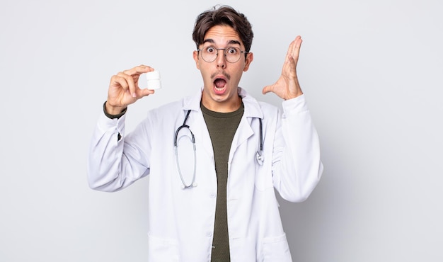 Young hispanic man screaming with hands up in the air. physician with pills bottle concept
