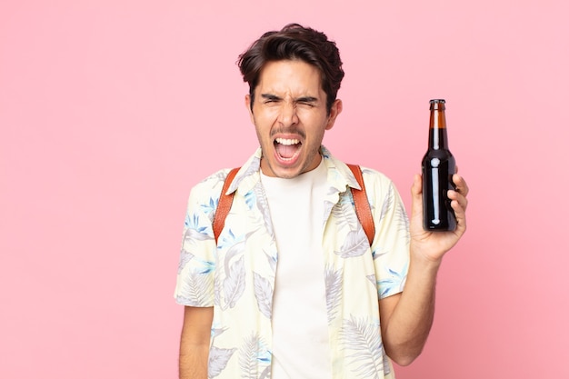 Young hispanic man shouting aggressively, looking very angry and holding a bottle of beer