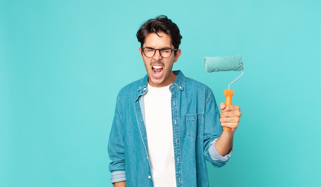 Young hispanic man shouting aggressively, looking very angry and holding a paint roller