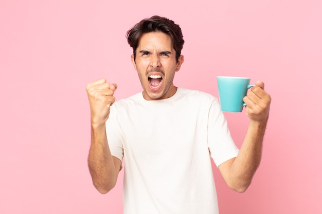 Young hispanic man shouting aggressively with an angry expression and holding a coffee mug