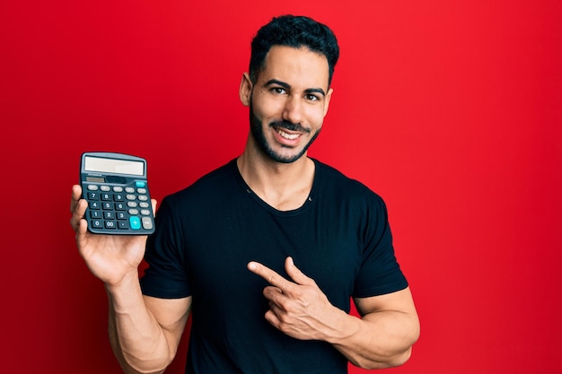 Young hispanic man showing calculator device smiling happy pointing with hand and finger