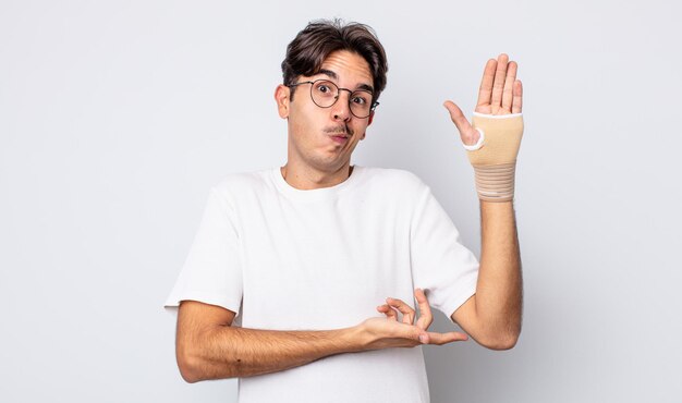 Young hispanic man shrugging, feeling confused and uncertain. hand bandage concept