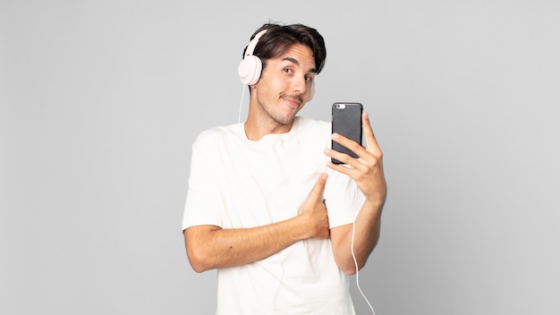 young hispanic man shrugging, feeling confused and uncertain with headphones and smartphone