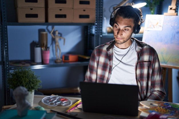 Young hispanic man sitting at art studio with laptop late at night smiling looking to the side and staring away thinking.