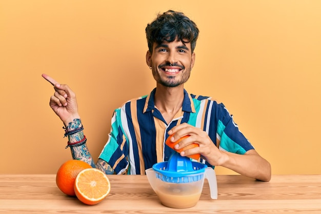 Young hispanic man sitting on the table using juicer smiling happy pointing with hand and finger to the side