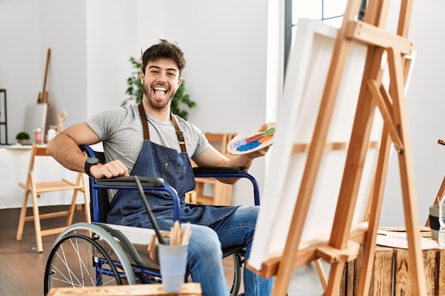 Молодой латиноамериканец сидит на инвалидной коляске и рисует в художественной студии, высунув язык, довольный забавной концепцией эмоций выражения