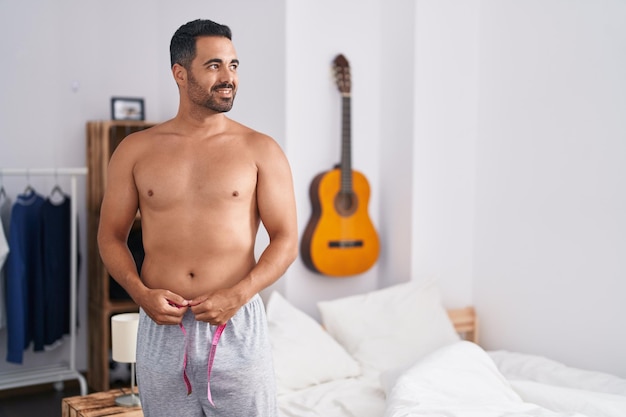 Young hispanic man smiling confident measuring waist at bedroom