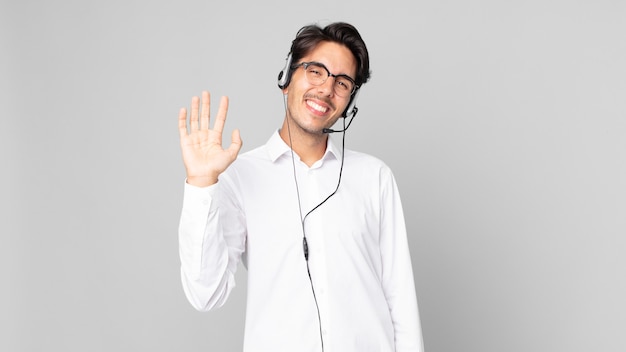 young hispanic man smiling happily, waving hand, welcoming and greeting you. telemarketer concept