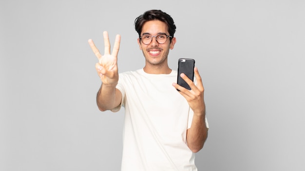 Young hispanic man smiling and looking friendly, showing number three and holding a smartphone