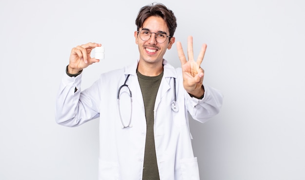 Young hispanic man smiling and looking friendly, showing number three. physician with pills bottle concept