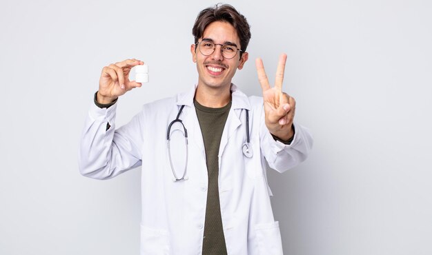 Young hispanic man smiling and looking friendly, showing number two. physician with pills bottle concept