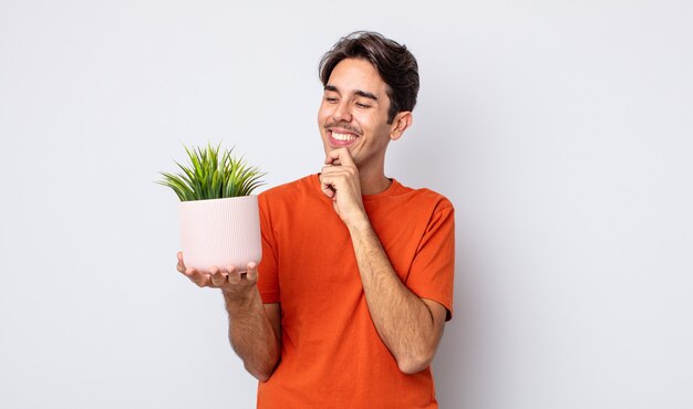 young hispanic man smiling with a happy, confident expression with hand on chin. decorative plant concept