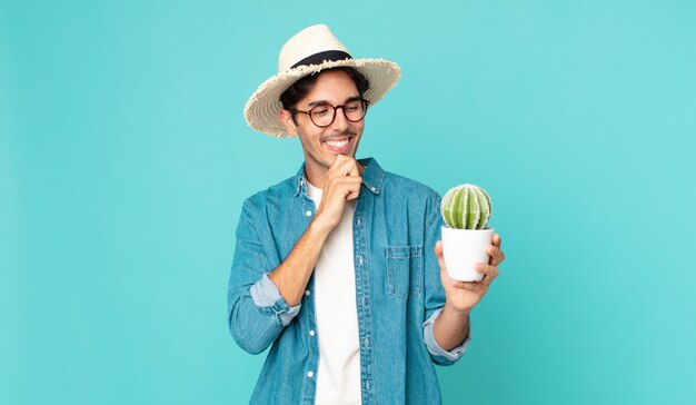 Young hispanic man smiling with a happy, confident expression with hand on chin and holding a cactus