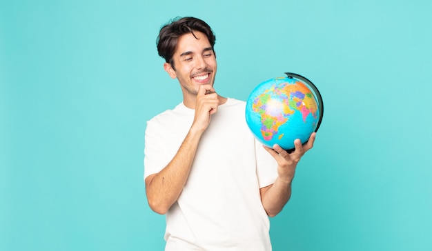 Young hispanic man smiling with a happy, confident expression with hand on chin and holding a world globe map