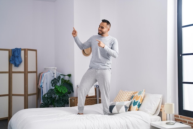 Young hispanic man standing on bed dancing at bedroom