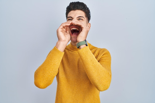 Young hispanic man standing over blue background shouting angry out loud with hands over mouth