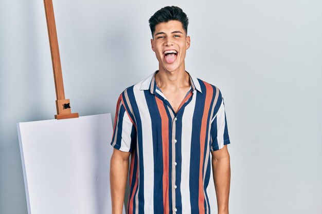 Young hispanic man standing close to empty canvas sticking tongue out happy with funny expression. emotion concept.