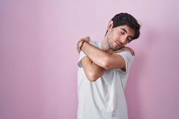 Young hispanic man standing over pink background hugging oneself happy and positive smiling confident self love and self care