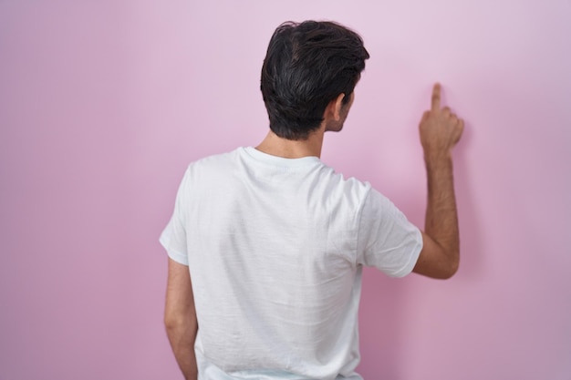 Young hispanic man standing over pink background posing backwards pointing ahead with finger hand