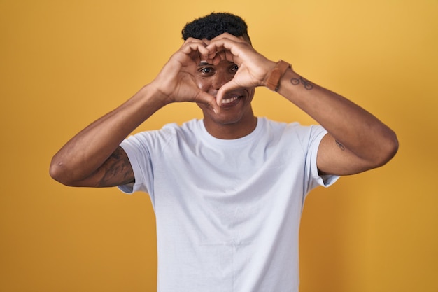 Young hispanic man standing over yellow background doing heart shape with hand and fingers smiling looking through sign