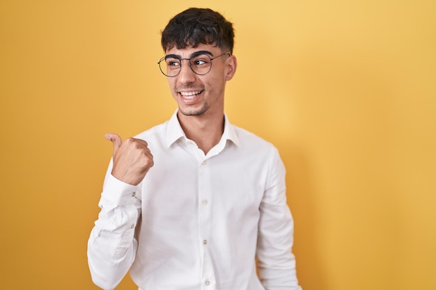 Young hispanic man standing over yellow background smiling with happy face looking and pointing to the side with thumb up