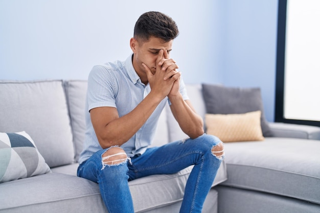 Молодой испаноязычный мужчина испытывает стресс, сидя дома на диване.