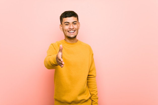 Young hispanic man stretching hand at camera in greeting gesture.