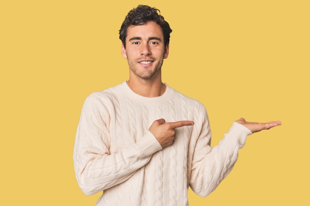 Young Hispanic man in studio excited holding a copy space on palm