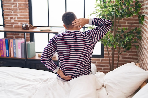 Young hispanic man suffering for back injury sitting on bed at bedroom