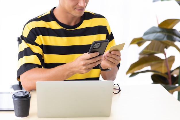 Young hispanic man using credit card to payment online with smartphone and laptop computer at home or creative workplace office.