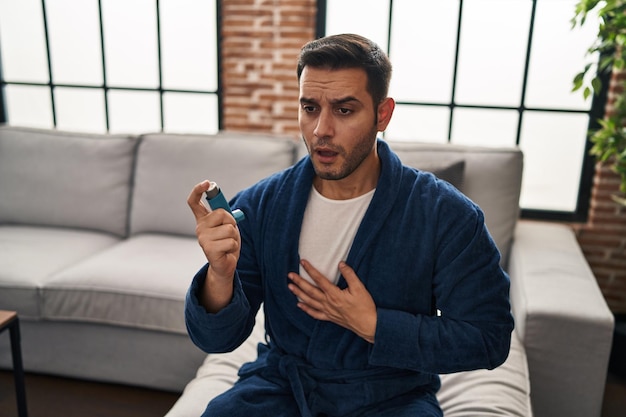 Young hispanic man using inhaler sitting on sofa at home