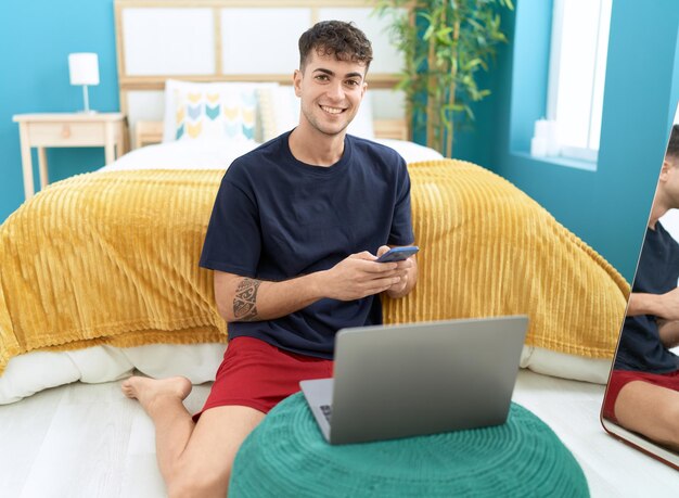 Photo young hispanic man using laptop and smartphone sitting on floor at bedroom