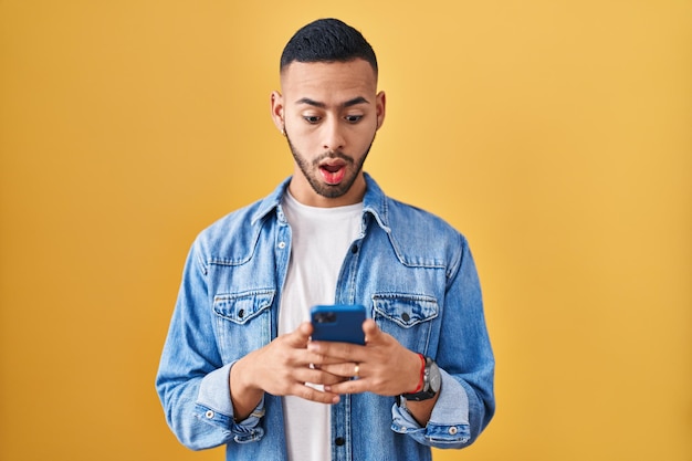 Young hispanic man using smartphone typing message in shock face, looking skeptical and sarcastic, surprised with open mouth