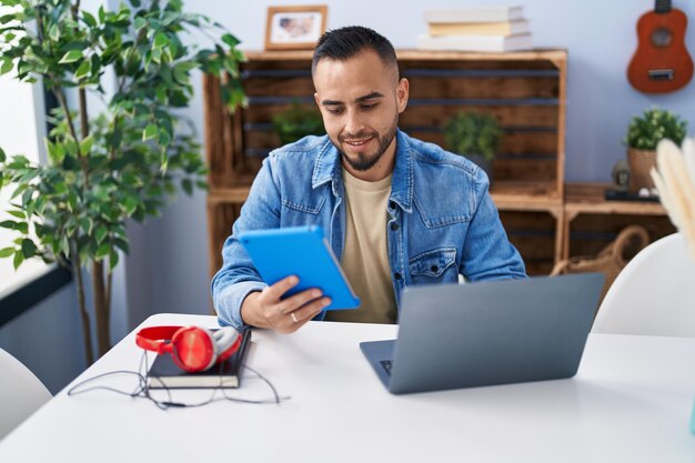 Young hispanic man using touchpad and laptop sitting on table at home