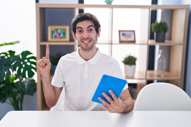 Young hispanic man using touchpad sitting on the table smiling with an idea or question pointing finger with happy face, number one