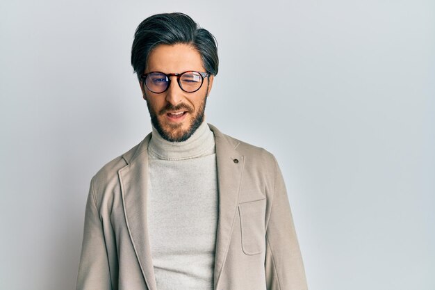 Young hispanic man wearing business jacket and glasses winking looking at the camera with sexy expression cheerful and happy face