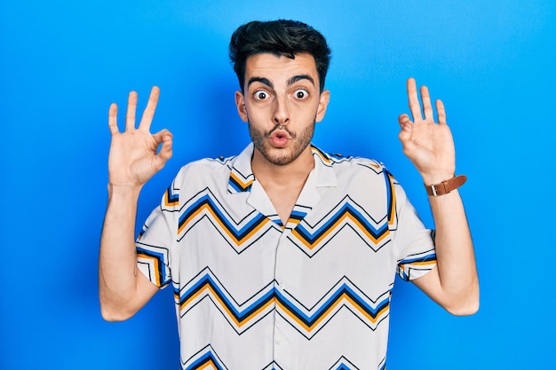 Young hispanic man wearing casual clothes looking surprised and shocked doing ok approval symbol with fingers. crazy expression