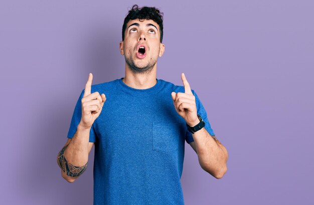 Young hispanic man wearing casual t shirt amazed and surprised looking up and pointing with fingers and raised arms