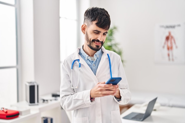 Young hispanic man wearing doctor uniform using smartphone at clinic