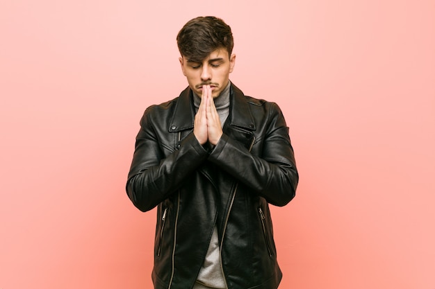 Young hispanic man wearing a leather jacket holding hands in pray near mouth, feels confident.