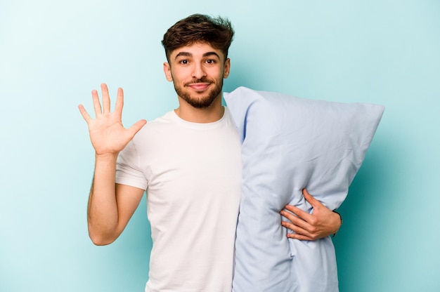 Молодой латиноамериканец в пижаме держит подушку на синем фоне, весело улыбаясь, показывая пальцами номер пять