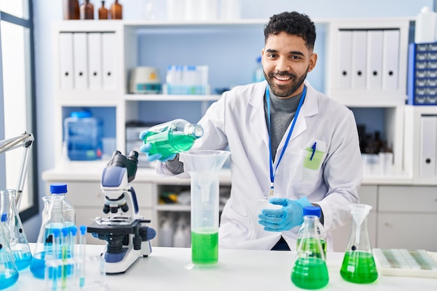 Young hispanic man wearing scientist uniform measuring liquid at laboratory
