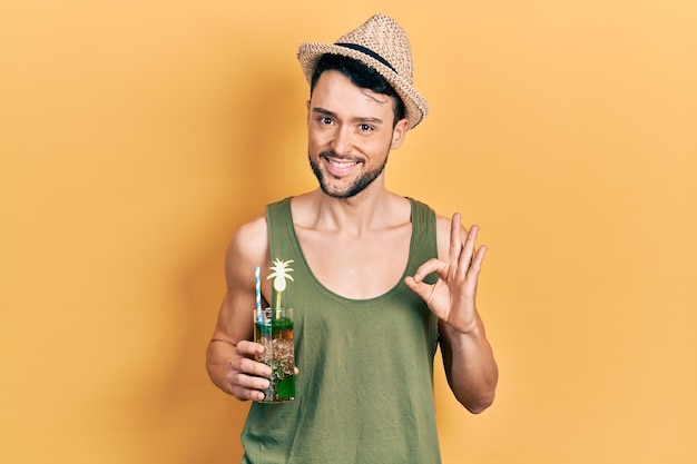 Молодой латиноамериканец в летней шляпе пьет мохито, делает знак "ок" с пальцами, дружелюбно улыбаясь, жестикулируя отличный символ